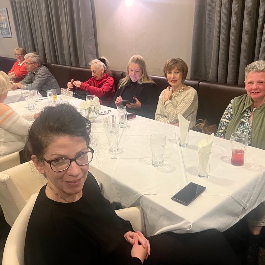 Mehrere Menschen sitzen in einem Restaurant um einen Tisch herum, lächeln und unterhalten sich. Der Tisch ist mit einer weißen Tischdecke bedeckt und hat Gläser, Teller und ein Handy. Im Hintergrund ist eine Wand mit einem Bilderrahmen und Vorhängen zu sehen.