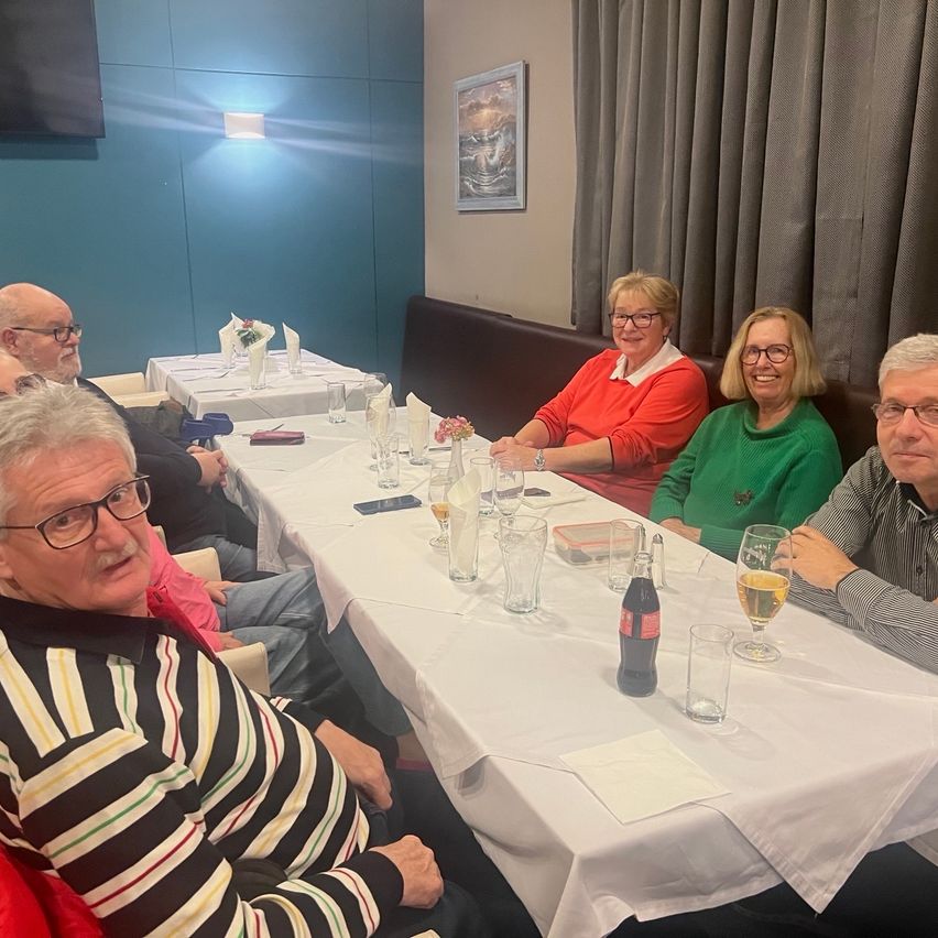 Mehrere Personen sitzen um einen Tisch in einem Restaurant. Gläser, Flaschen und Servietten liegen auf dem Tisch. Der Raum hat eine blaue Wand mit einem Bilderrahmen. Die Menschen lächeln.