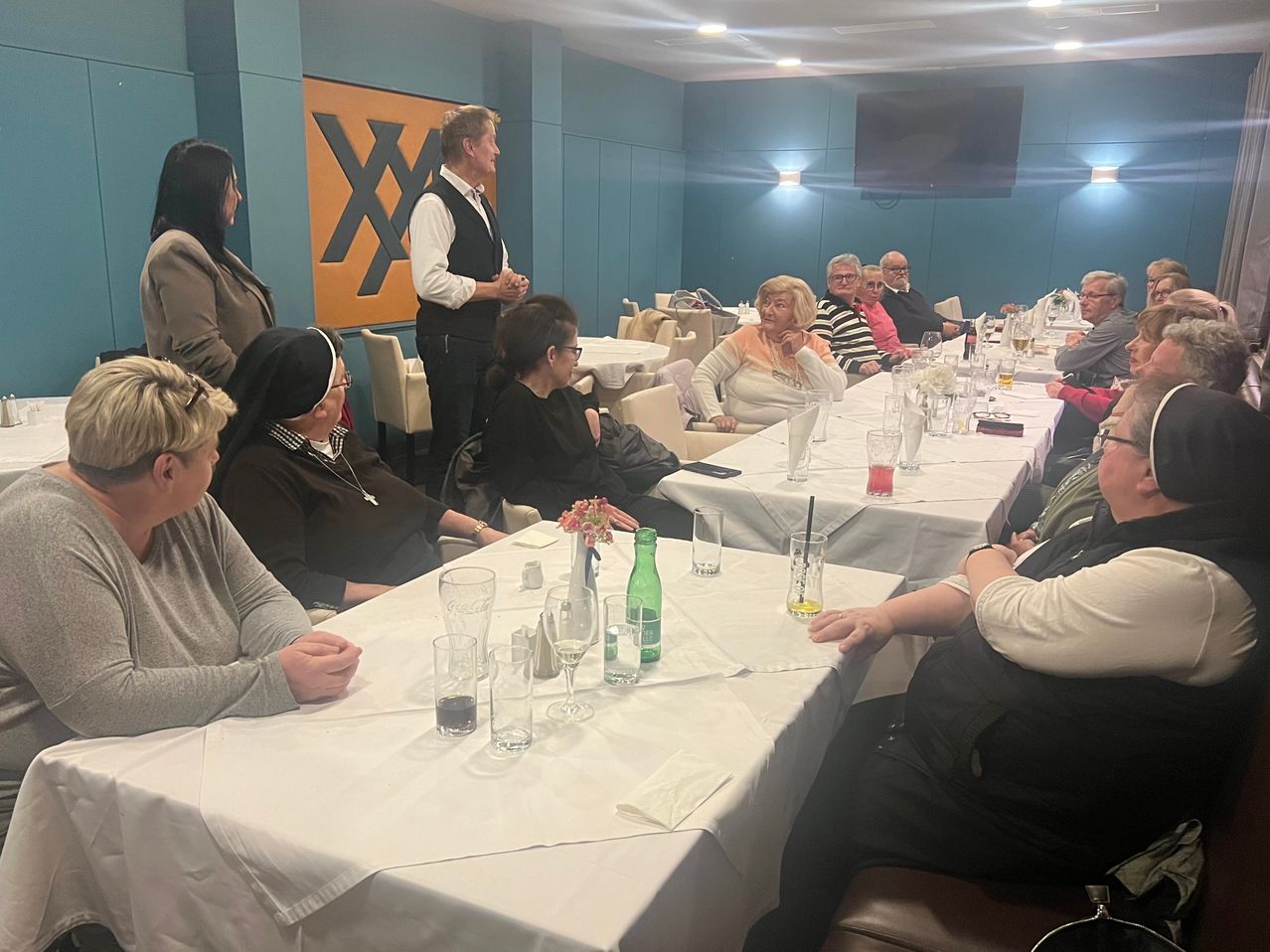 Eine Gruppe älterer Personen sitzt an einem langen Tisch in einem Restaurant, mit einer Frau vor ihnen. Der Tisch ist mit Gläsern, Flaschen und Servietten gedeckt. Ein großer gelber Buchstabe 'X' befindet sich an der Wand.