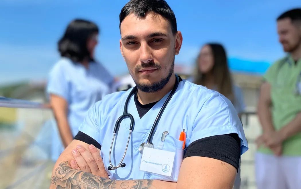 Ein Krankenpfleger mit einem Stethoskop um den Hals steht mit verschränkten Armen und trägt ein hellblaues Scrub-Oberteil. Hinter ihm sind zwei Krankenschwestern in ähnlichen Uniformen zu sehen.