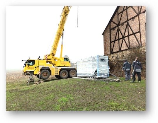 Ein gelber Kran steht auf einem Grasfeld und hebt einen großen, abgedeckten Container. Zwei Personen in Jacken stehen in der Nähe, mit einem Backsteingebäude im Hintergrund.