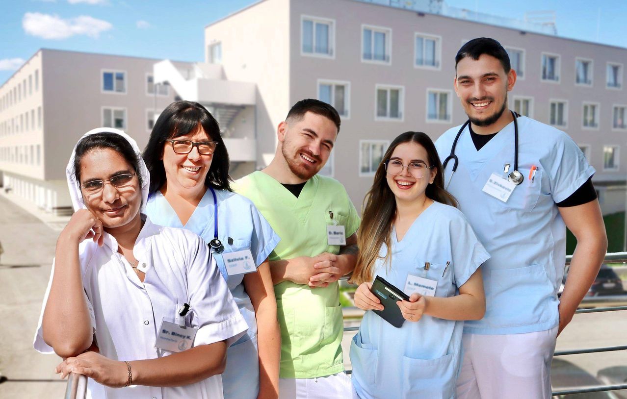Fünf Gesundheitsfachkräfte, darunter zwei Frauen und drei Männer, lächeln für die Kamera vor einem Gebäude. Sie tragen blaue Uniformen und einer hält einen Geldbeutel.