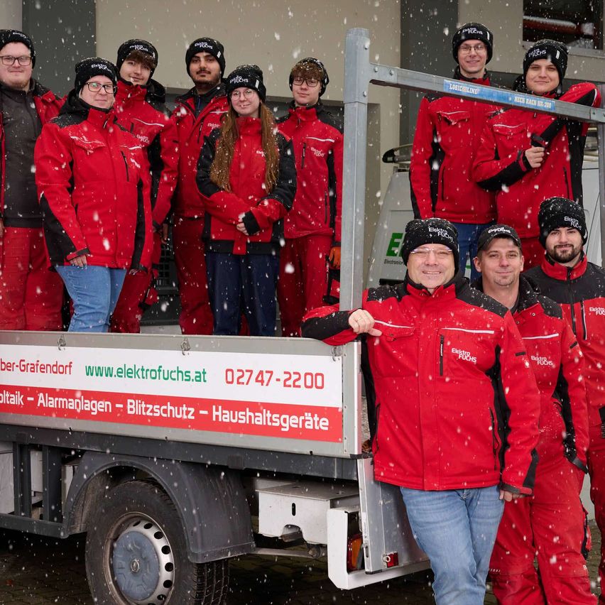 Eine Gruppe von Elektrikern in roten Jacken steht vor einem Lastwagen unter schneereichen Bedingungen. Der Lastwagen hat Text im Zusammenhang mit elektrischen Dienstleistungen.