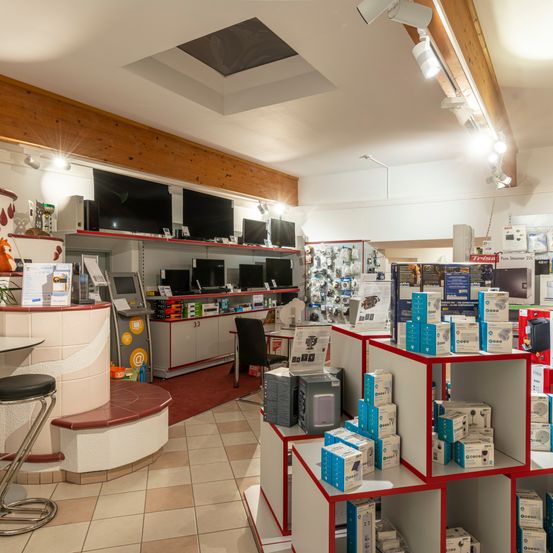 Ein Elektronikgeschäft mit gefliesten Böden, Regalen mit Kisten und Monitoren im Schaufenster. Eine Theke mit einem Hocker und einem roten Teppich auf dem Boden.
