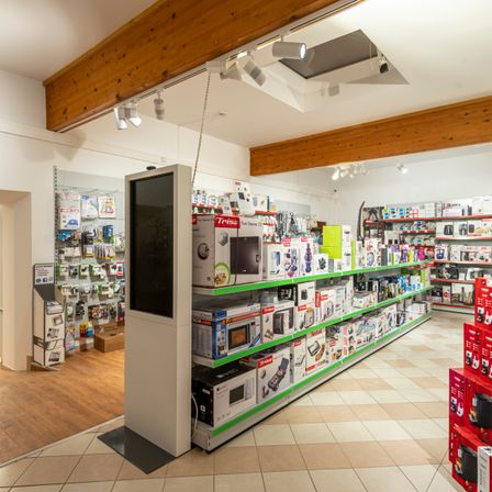 Ein Elektronikgeschäft mit hölzernen Deckenbalken und einem gefliesten Boden. Eine zentrale Ausstellung zeigt verschiedene Küchengeräte, darunter Mikrowellen und Toaster. Produkte sind ordentlich auf Regalen mit grünen Rändern angeordnet.