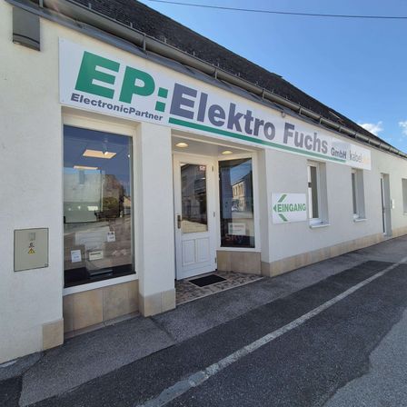 Vorderansicht eines weißen Gebäudes mit einem grünen Schild, das EP: Elektro Fuchs steht. Es gibt eine Glastür und Fenster mit der Reflexion eines Gebäudes und des Himmels.