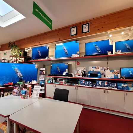 Ein Elektronikgeschäft mit mehreren großen Bildschirmen, die Schiffe anzeigen. Die Bildschirme sind an der Wand montiert. Die darunterliegenden Regale sind mit verschiedenen elektronischen Produkten gefüllt. Vorne stehen zwei Tische.