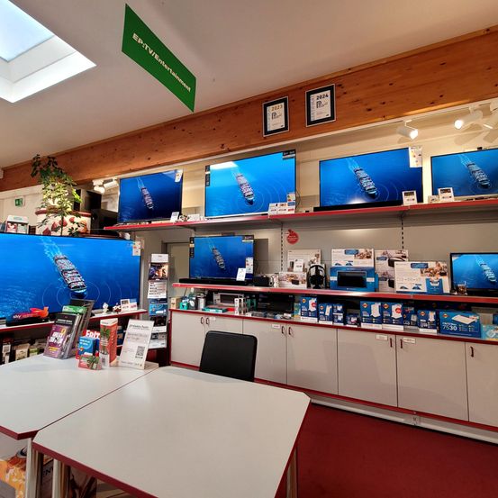 Ein Elektronikgeschäft mit mehreren großen Bildschirmen, die Schiffe anzeigen. Die Bildschirme sind an der Wand montiert. Die darunterliegenden Regale sind mit verschiedenen elektronischen Produkten gefüllt. Vorne stehen zwei Tische.