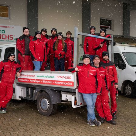 Eine Gruppe von Elektrikern in roten Uniformen posiert für ein Foto vor einem LKW bei Schneewetter. Sie stehen auf der Rückseite des LKW.