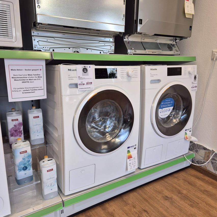 Zwei weiße Waschmaschinen sind auf einem Holzboden ausgestellt, mit einer weißen Wand im Hintergrund. Die Waschmaschinen haben einen Aufkleber mit der Aufschrift 'Miele' und ein Schild mit der Aufschrift 'Miele Aktion - Sauberer TwinDos.' Auf der linken Seite befinden sich Flaschen mit Reinigungsmittel.