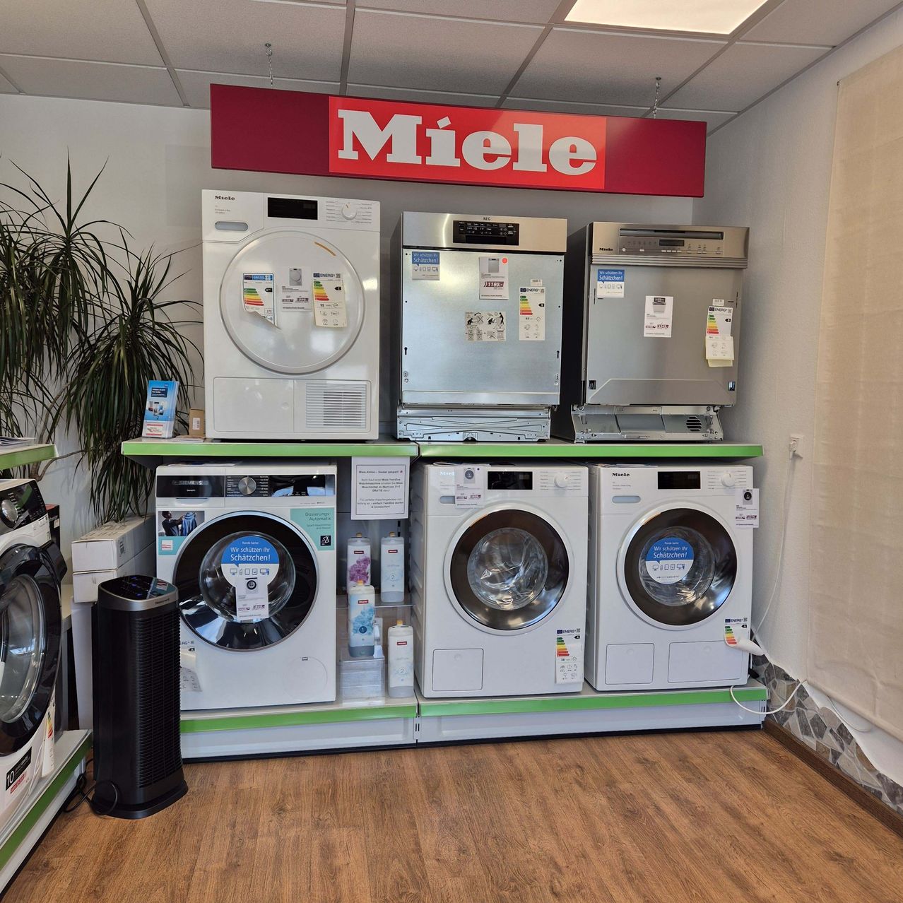 Ein Elektrogeräteladen mit einem Miele-Schild an der Decke. Verschiedene weiße Geräte sind auf Regalen ausgestellt, darunter Waschmaschinen, Trockner und Geschirrspüler.