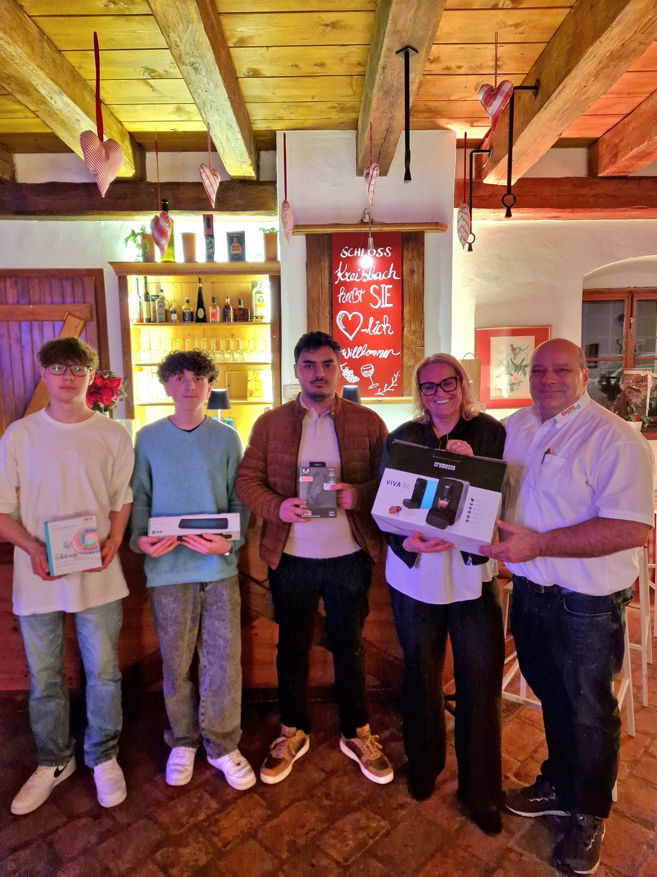 Vier Personen stehen zusammen und posieren für ein Foto. Die beiden Männer an den Seiten halten Geschenke. Die zentrale Person hält eine Kaffeemaschine. Hinter ihnen befindet sich ein Regal mit Spirituosenflaschen und einem Schild, das 'Schloss Kleinbach' besagt.