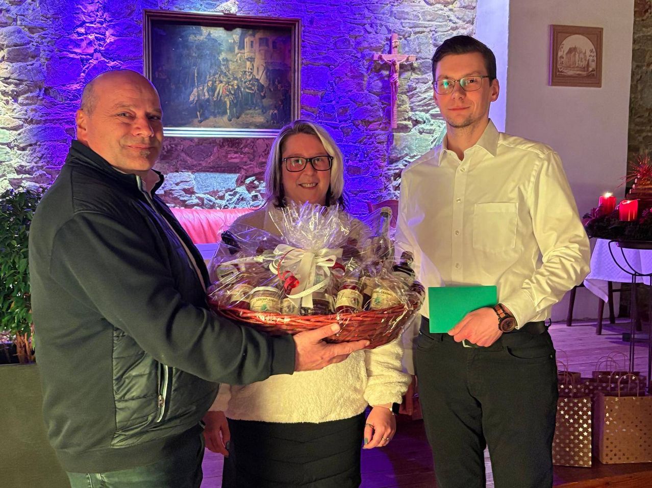 Drei Personen stehen in einem Raum, wobei eine Frau einen Korb mit Lebensmitteln hält. Hinter ihnen ist ein Kreuz an der Wand befestigt.