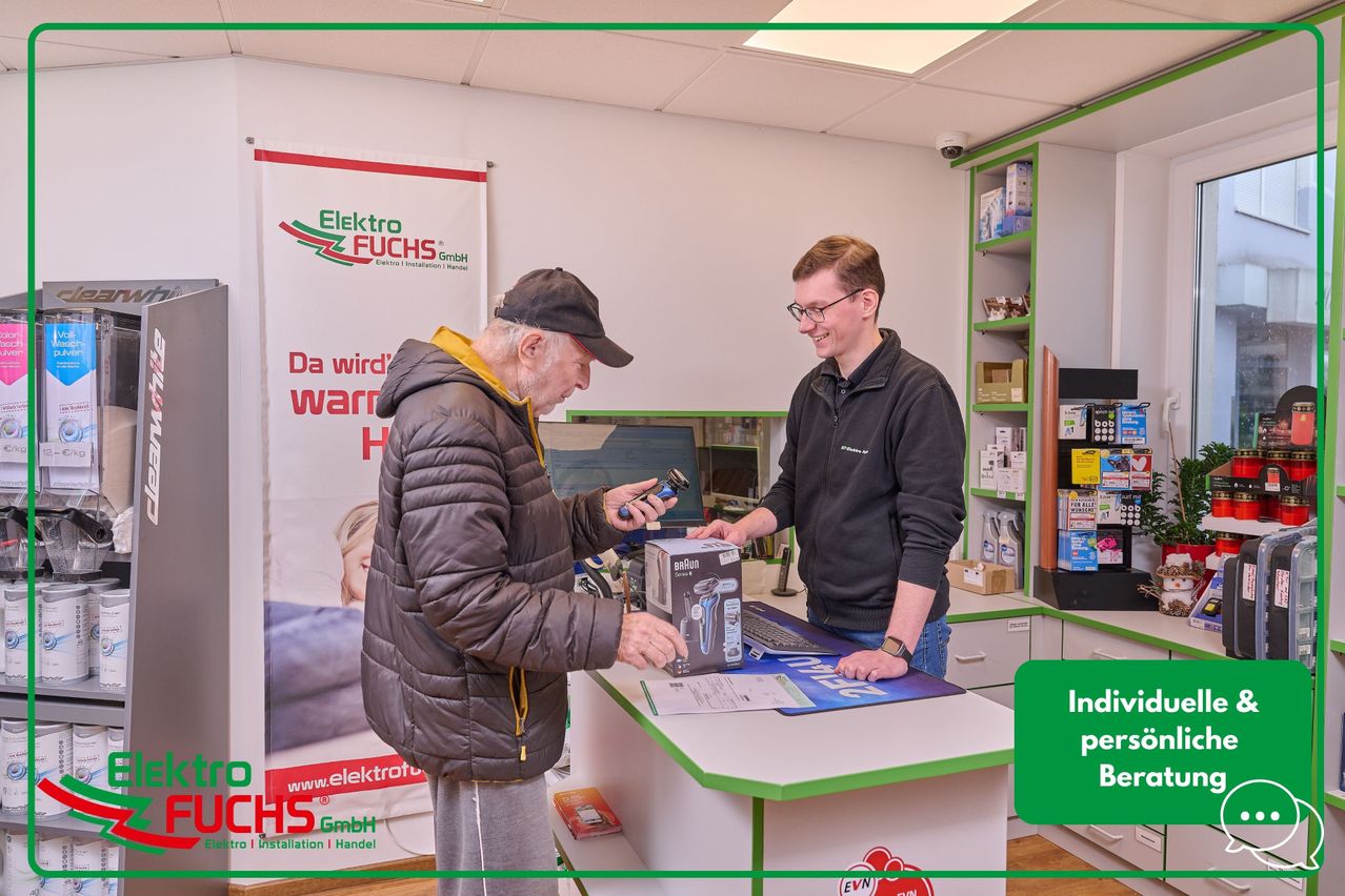 Zwei Männer stehen an einem Tresen in einem Elektronikgeschäft. Ein älterer Mann hält einen Braun Rasierer, während ein jüngerer Mann zusieht. Im Hintergrund befindet sich ein Banner mit den Worten 'Da wird' warr' und dem Logo von Elektro Fuchs.
