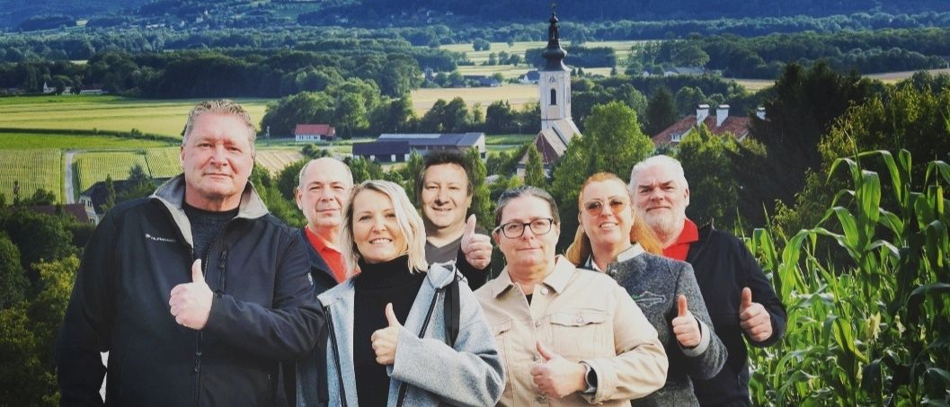 Eine Gruppe von Menschen posiert für ein Foto vor einer Kirche und einer malerischen Kulisse. Sie alle zeigen Daumen hoch.