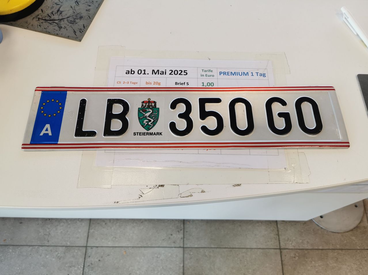 Ein Nummernschild mit der Aufschrift 'LB 350 GO' ist auf einer weißen Oberfläche zu sehen. Links davon befindet sich eine blau-gelbe EU-Flagge. Ein Preisschild mit Tarif trägt die Aufschrift 'PREMIUM 1 Tag' und ist vom 1. Mai 2025 datiert.