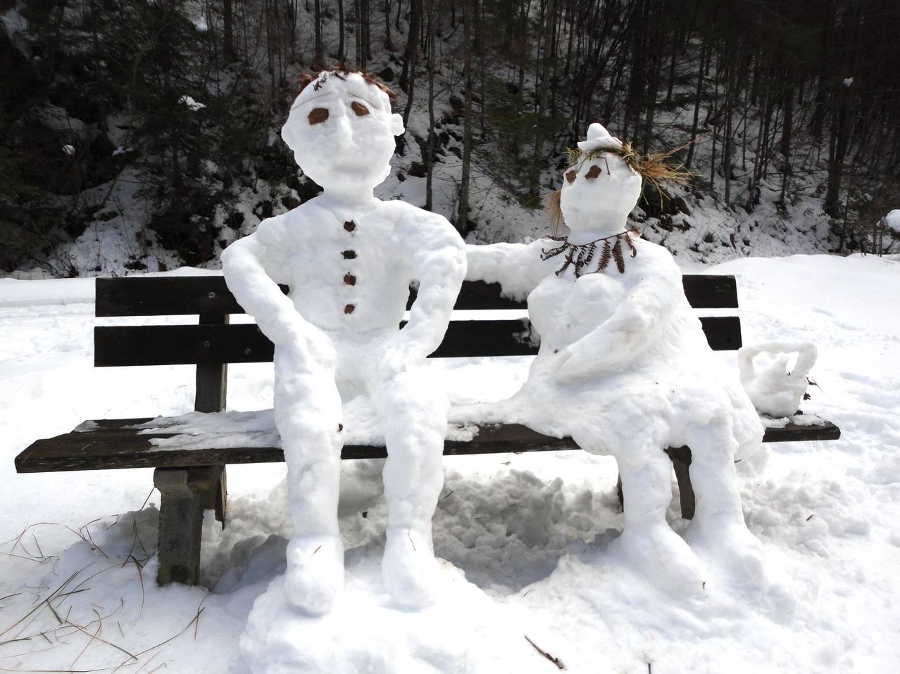 Zwei Schneemänner sitzen auf einer Bank in einer verschneiten Landschaft mit Bäumen und schneebedecktem Boden. Der Schneemann auf der linken Seite hat einen traurigen Gesichtsausdruck und der rechte hat eine Krone auf dem Kopf.