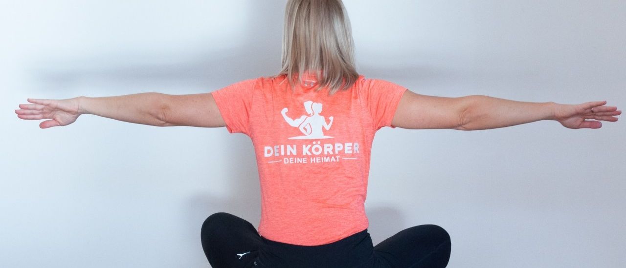 Eine Frau praktiziert Yoga auf einer Matte und trägt ein T-Shirt mit der Aufschrift 'Dein Körper Deine Heimat' und schwarze Leggings. Sie sitzt mit gekreuzten Beinen, ausgestreckten Armen und blickt nach vorne. Der Hintergrund ist eine schlichte weiße Wand.
