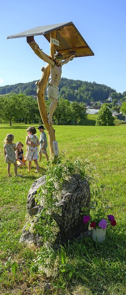 Kinder versammeln sich um ein Holzkreuz mit einer Figur von Jesus. Das Kreuz steht auf einer Rasenfläche mit einem großen Felsen und Blumen in der Nähe.