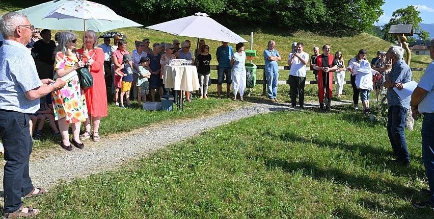 Eine Zusammenkunft im Freien mit vielen stehenden Personen, einige mit Regenschirmen. Ein Tisch mit weißem Tuch ist aufgestellt. Der Bereich ist von Grün umgeben.