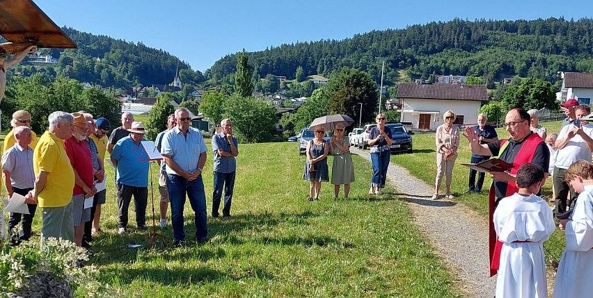 Eine Gruppe von Menschen versammelt sich auf einem grünen Feld unter klarem Himmel, während ein Priester aus einem Buch liest. Autos und Häuser sind im Hintergrund zu sehen.