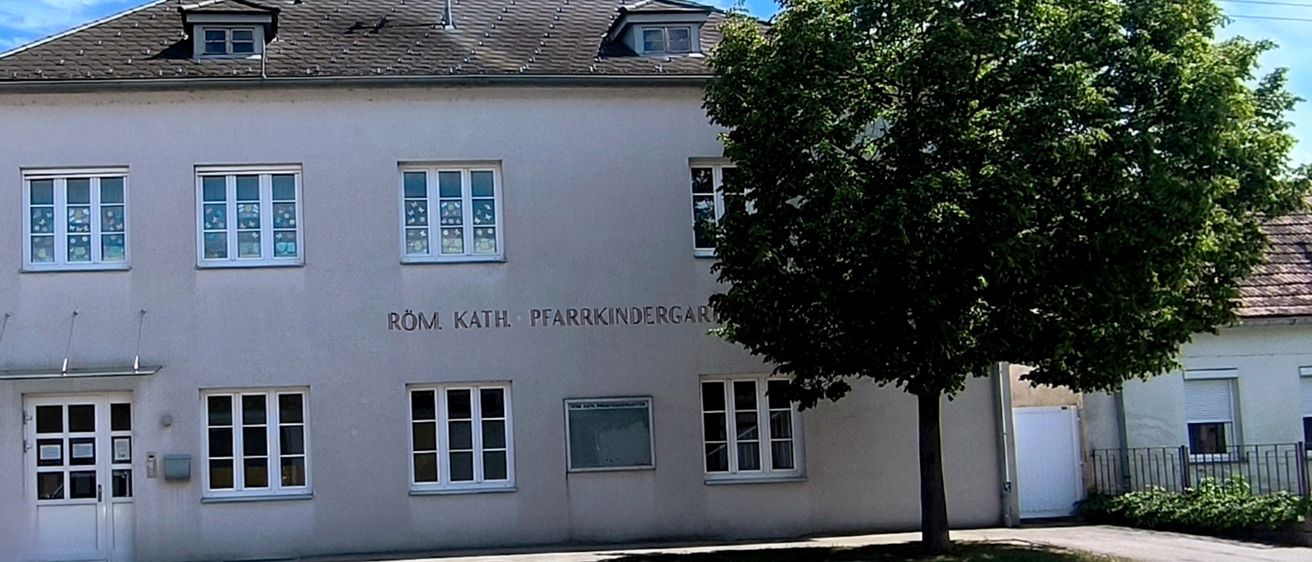 Ein weißes Gebäude mit einem braunen Dach hat mehrere Fenster. Es trägt den Namen Röm. Kath. Pfarrkindergarten. Ein Baum steht vor dem Gebäude. Der Himmel ist blau mit Wolken.