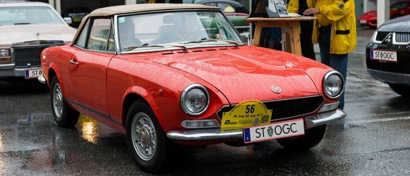 Ein roter Oldtimer mit einem gelben Nummernschild ist auf einer nassen Straße geparkt. In der Nähe stehen Leute mit Regenschirmen, einer spricht in ein Mikrofon.