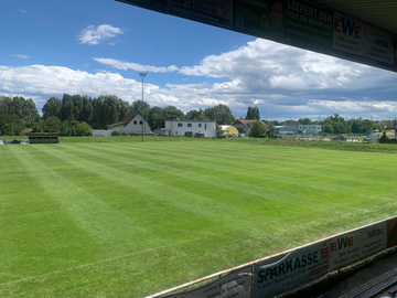 Ein leerer Sportplatz mit grünem Rasen unter einem teilweise bewölkten Himmel. Bäume sind in der Ferne zu sehen, und Gebäude sind sichtbar. Ein weißes Gebäude befindet sich in der Mitte, mit einer Straßenlaterne in der Mitte.