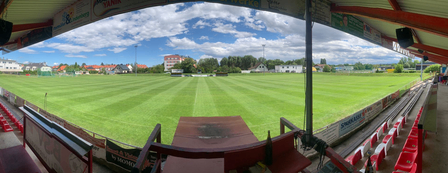 Ein Panoramablick auf ein leeres Stadion mit saftig grünem Rasen, roten Sitzen und einem offenen Dach. Der Himmel ist blau mit verstreuten Wolken.