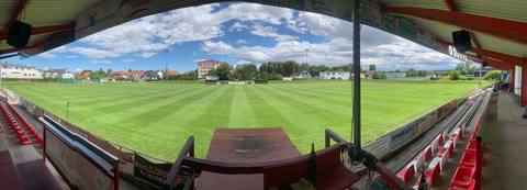 Ein Panoramablick auf ein leeres Stadion mit saftig grünem Rasen, roten Sitzen und einem offenen Dach. Der Himmel ist blau mit verstreuten Wolken.