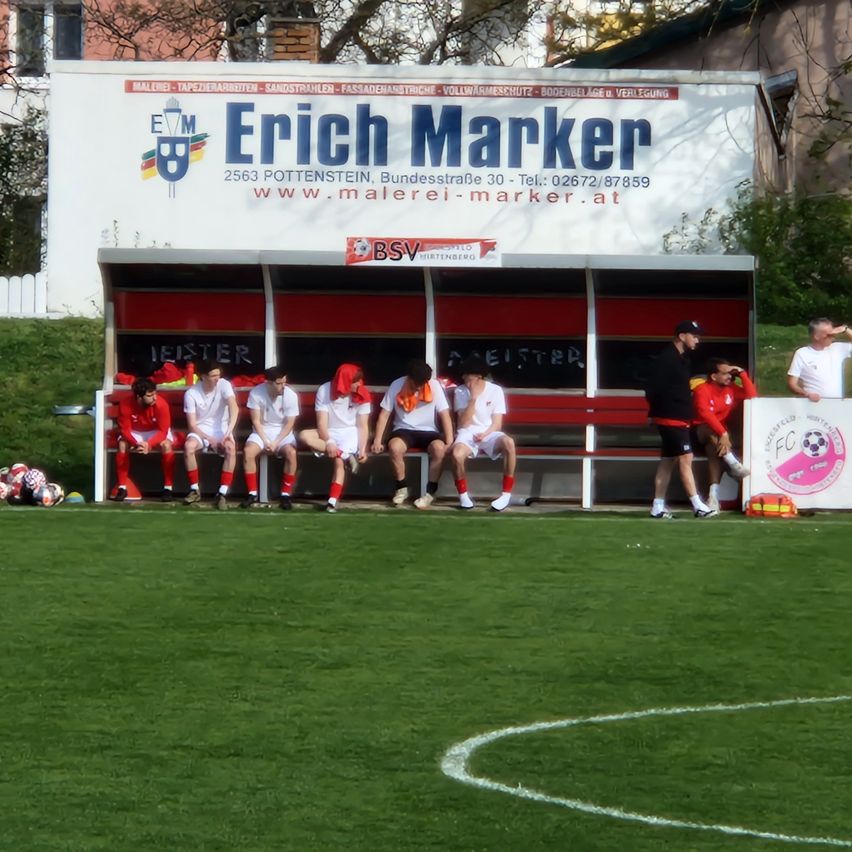 Spieler sitzen auf einer roten Bank auf einem Fußballfeld. Ein weißer Lieferwagen mit Erich Marker steht hinter ihnen.