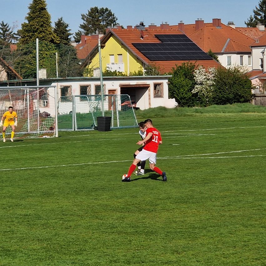 Zwei Fußballspieler spielen auf einem Feld mit einem Tor im Hintergrund. Ein Spieler in Rot tritt den Ball, während ein Spieler in Schwarz verteidigt.