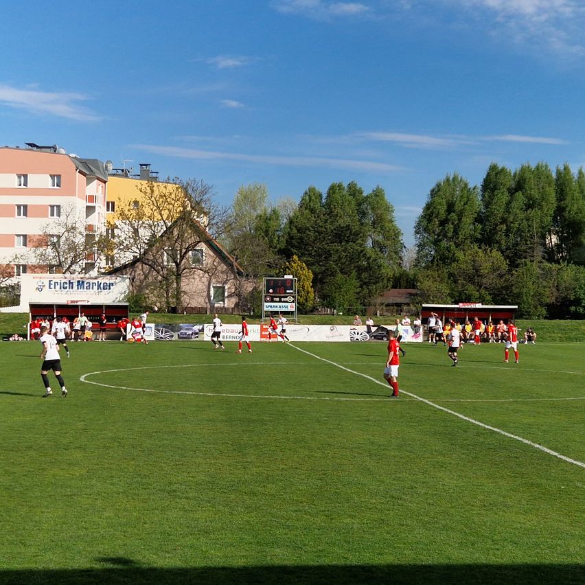 Ein Fußballspiel auf einem gut gepflegten Feld mit Spielern in roten und weißen Trikots. Ein Anzeigetafel ist im Hintergrund mit Gebäuden und Bäumen sichtbar.