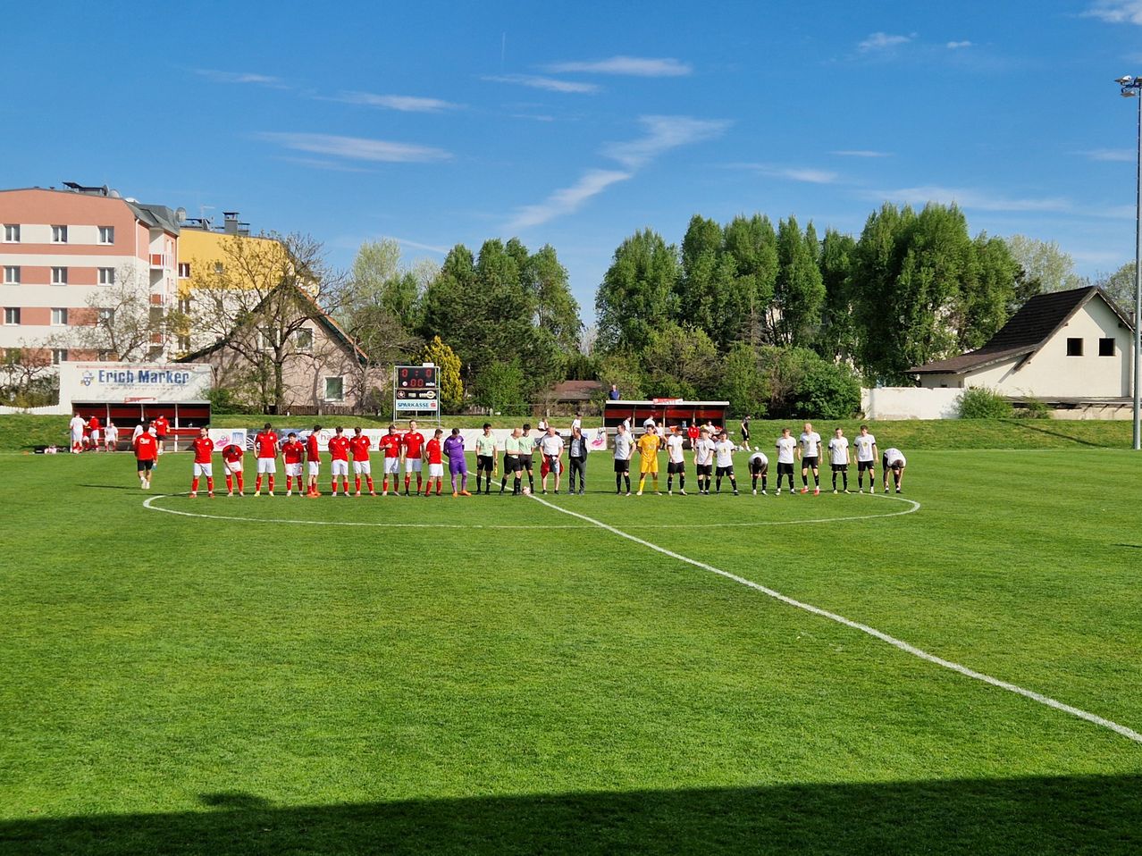 Eine Fußballmannschaft steht auf einem Rasenfeld. Dahinter befinden sich Gebäude und Bäume, und der Himmel ist blau mit einigen Wolken.