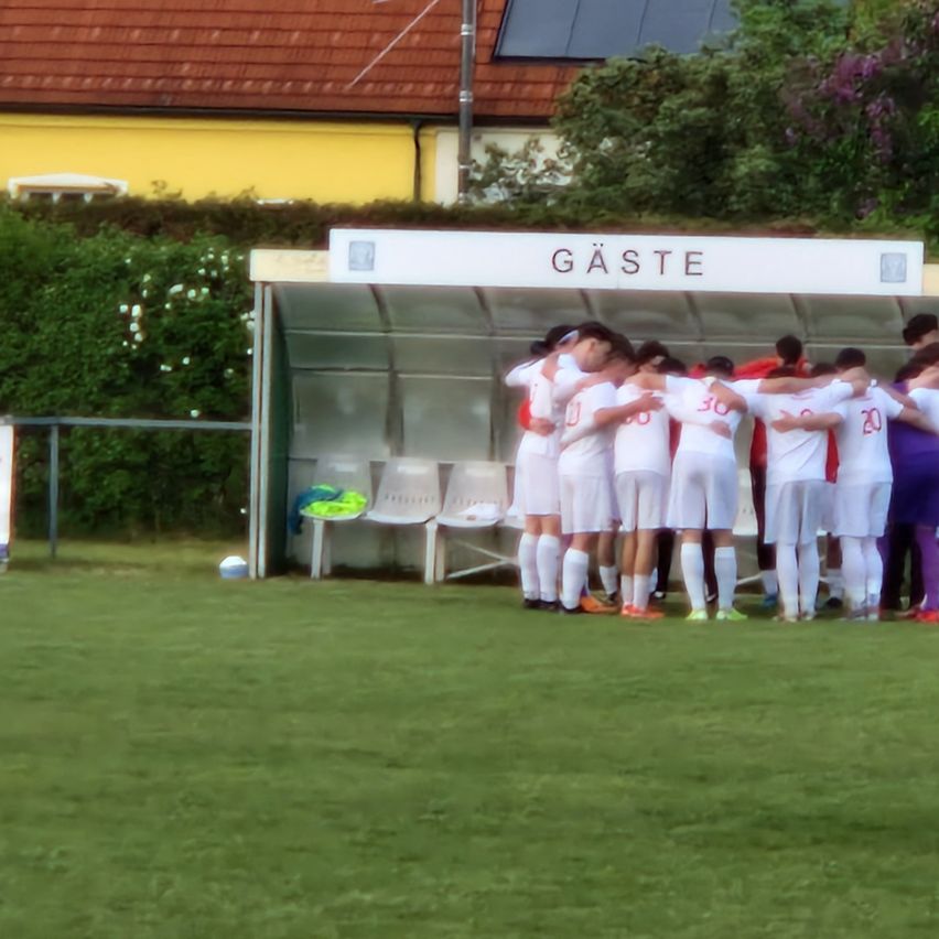 A team of young soccer players in white uniforms huddles together in a sports area. Behind them is a white shelter with the word 'GASTE' on top.