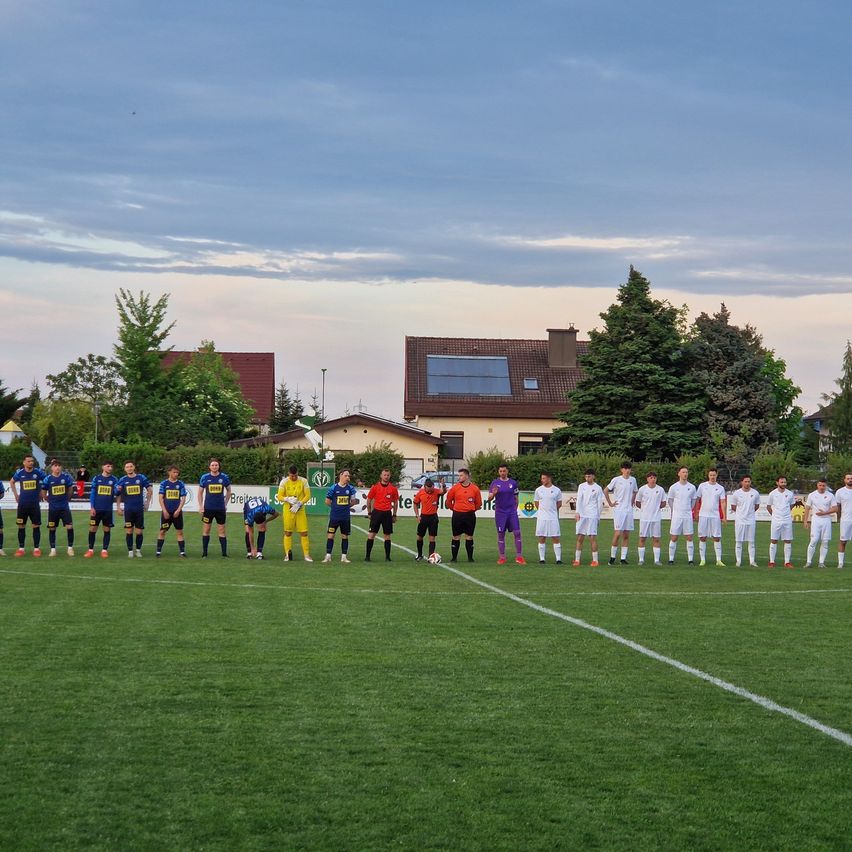 Two soccer teams in different colored jerseys stand on a field, facing each other, with houses and trees in the background.