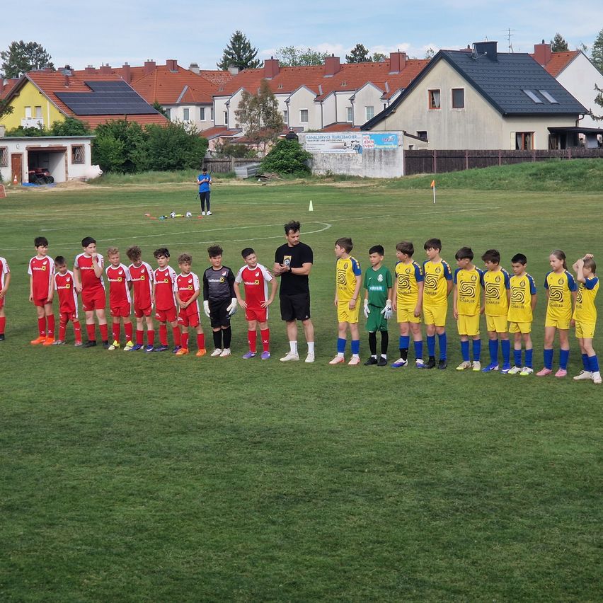Two youth soccer teams in red and yellow uniforms are lined up for a game. The coach in black stands between them. In the background, houses with solar panels are visible.