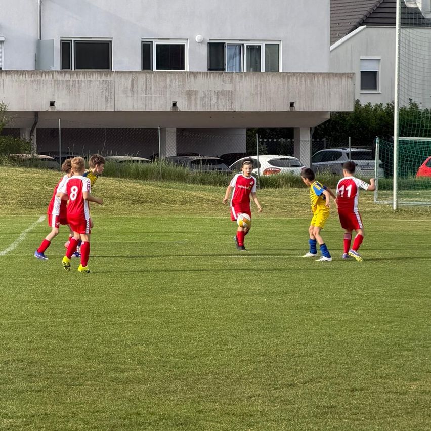 Young soccer players in red, yellow, and white uniforms are playing on a field, possibly in a competition. A building with several windows is in the background.