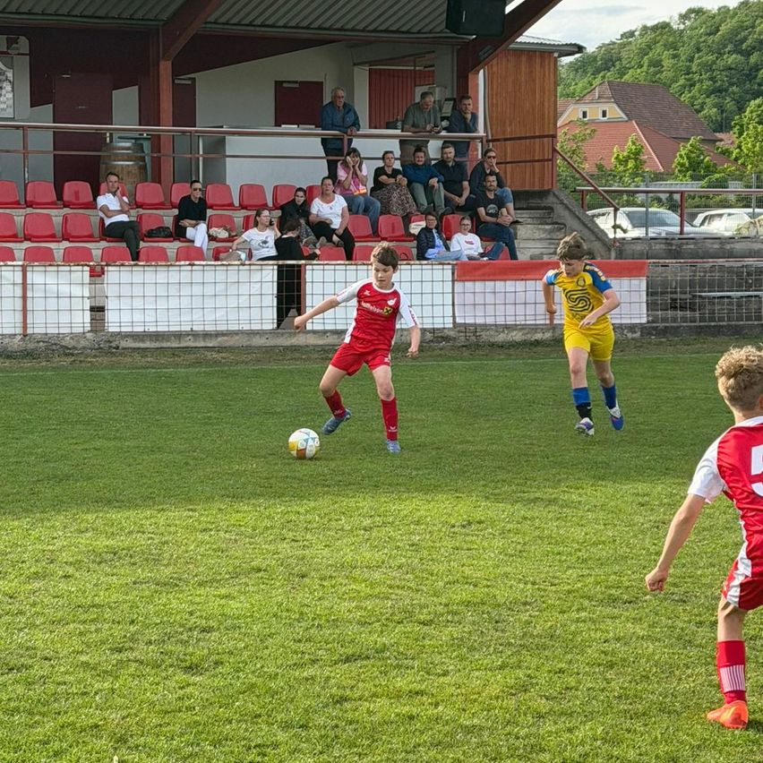 A soccer match in progress, players in red and yellow uniforms are chasing a ball. Spectators are seated in stands behind a fence. The field is green, and a red car is parked outside.