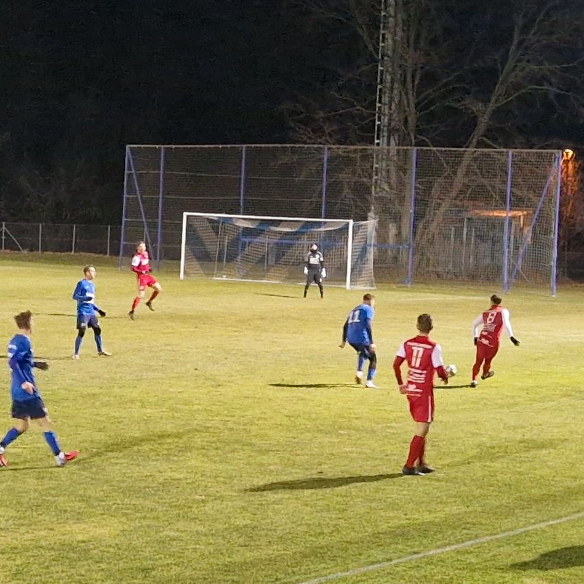 Ein Fußballspiel findet bei Nacht statt. Spieler in blauen und roten Trikots sind auf dem Feld und jagen dem Ball hinterher. Ein Torwart steht in der Nähe des Tores. Das Feld ist von einem Zaun umgeben.