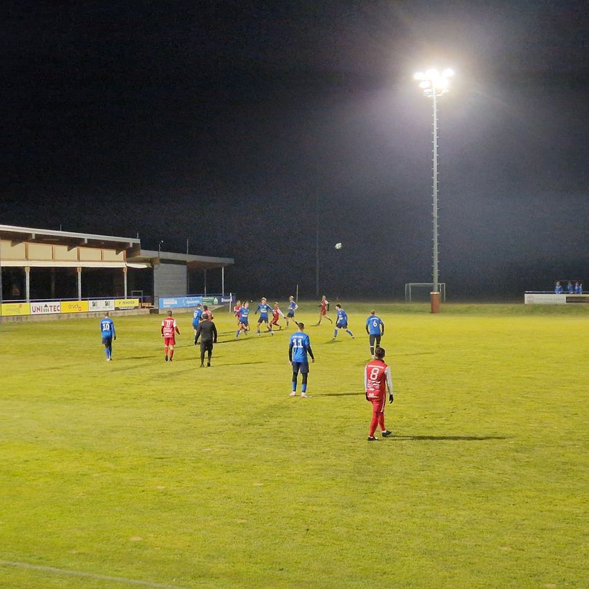 Ein nächtliches Fußballspiel findet statt, wobei Spieler in blauen und roten Trikots über das Feld verstreut sind. Helle Stadionlichter beleuchten den Bereich, und ein großes Tor ist im Hintergrund sichtbar.