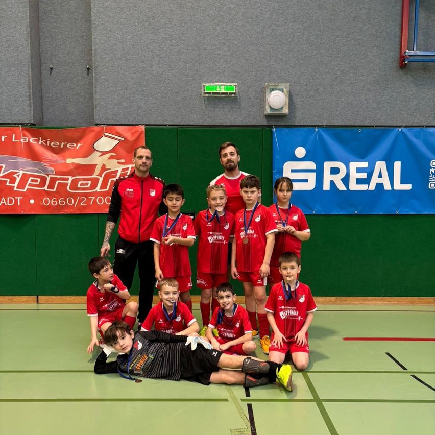 Eine Gruppe junger Jungen und Männer in roten Trikots mit Medaillen um den Hals, posiert für ein Foto auf einem Sportplatz. Hinter ihnen stehen zwei Männer in roten Uniformen und mehrere Banner hängen an einer grünen Wand.