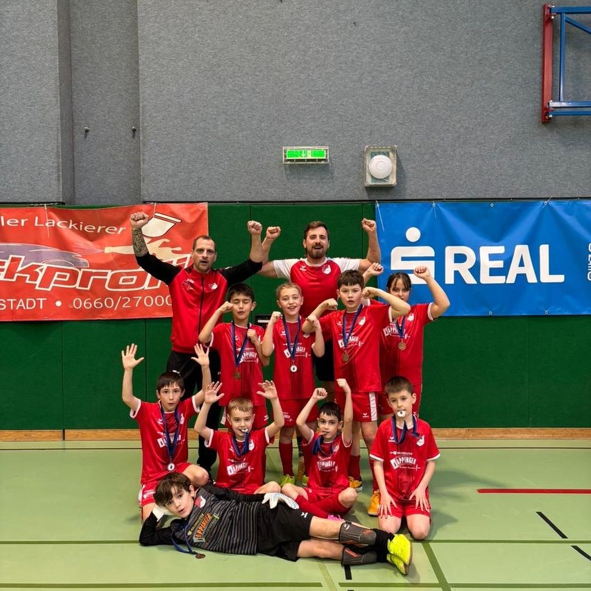 Eine Sportmannschaft posiert mit ihren Medaillen in einer Turnhalle. Zwei Trainer stehen hinter ihnen. Die Wand ist mit Bannern geschmückt.