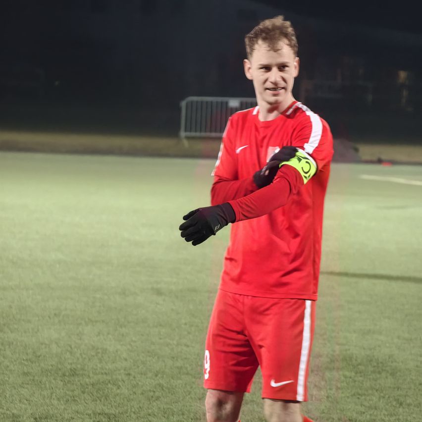 Ein Fußballspieler in einem roten Trikot mit weißen Streifen steht nachts auf einem Feld. Er lächelt und schaut in die Ferne.
