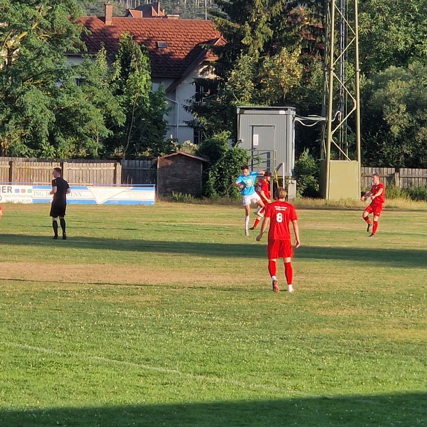Ein Fußballspiel auf einem Rasenfeld mit Spielern in roten und weißen Trikots. Bäume und ein Haus sind im Hintergrund zu sehen.