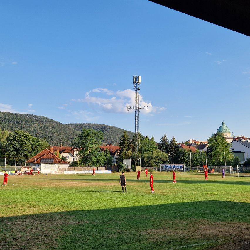 Eine Gruppe von Fußballspielern in roten Uniformen trainiert auf einem Feld mit einem Turm und Häusern im Hintergrund.