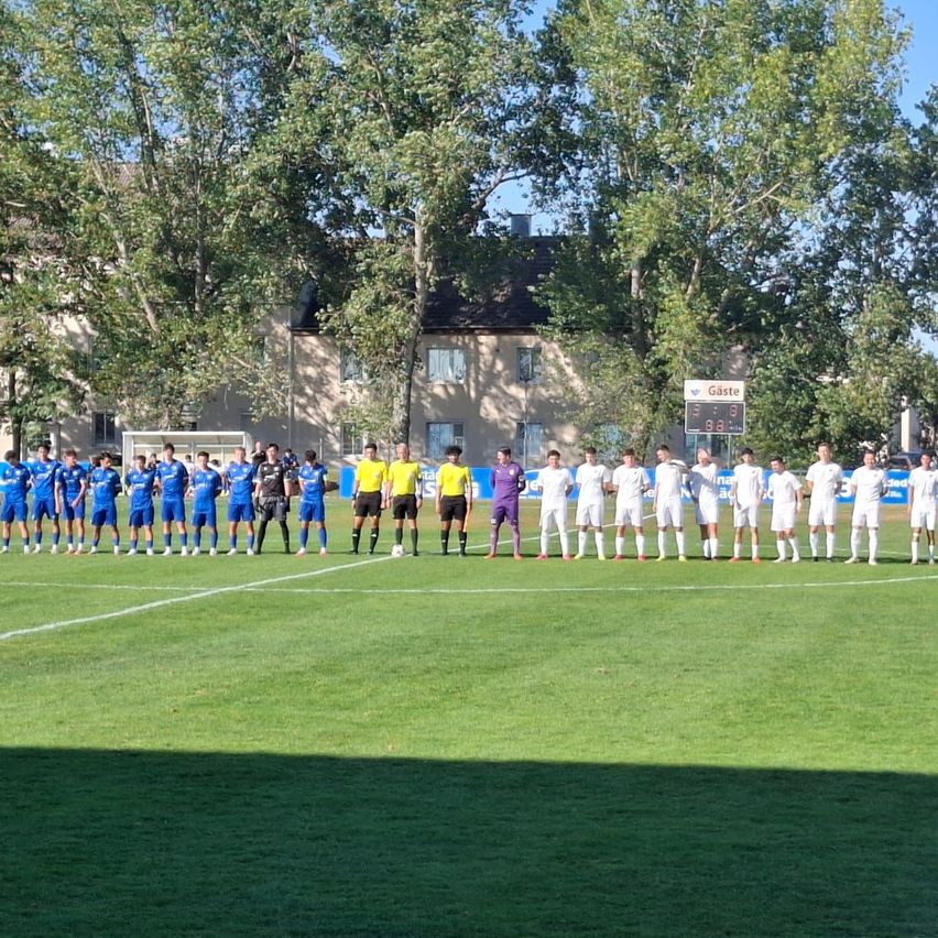Two soccer teams line up on a field with a scoreboard in the background. One team wears blue uniforms, and the other wears white.