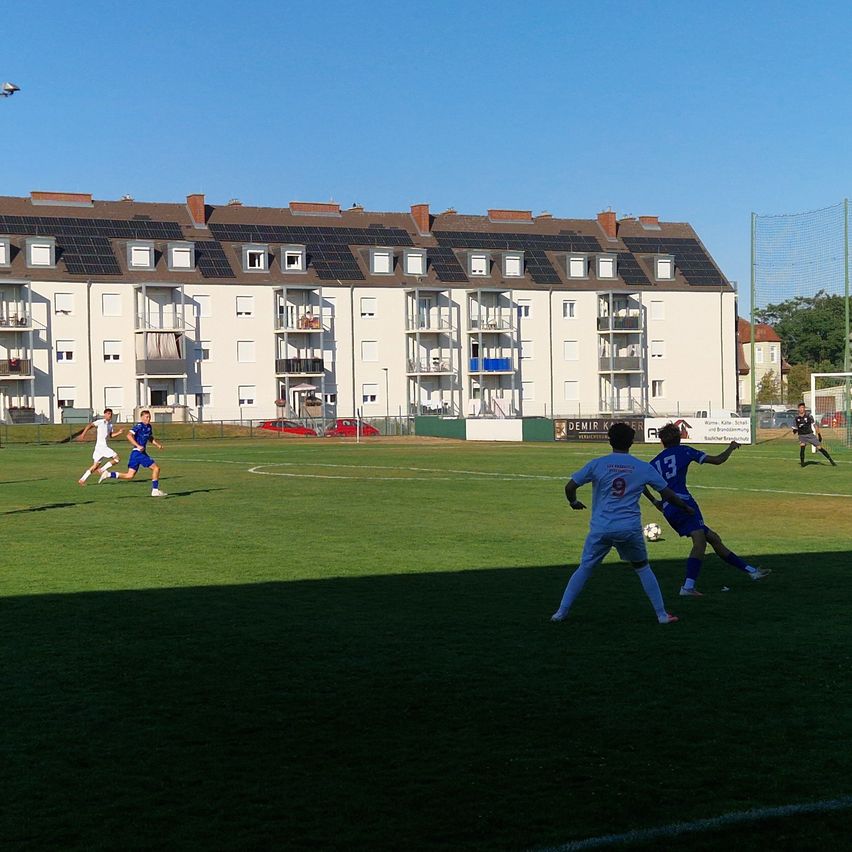 A soccer game is in progress on a field with players in white and blue uniforms. The field is surrounded by a fence, and in the background, there is a large building with solar panels on the roof.
