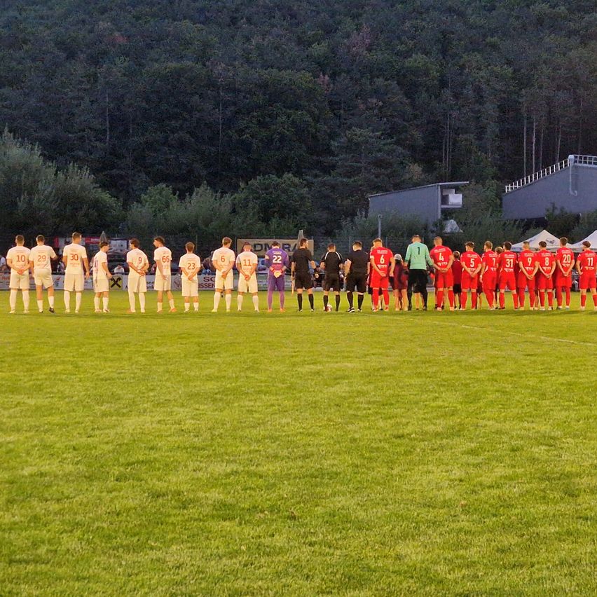 Eine Fußballmannschaft in roten Trikots steht in einer Reihe auf einem Feld, mit einer Kulisse aus Bäumen und einem Gebäude.