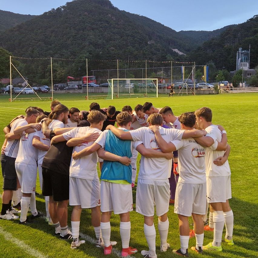 Eine Fußballmannschaft in weißen Trikots versammelt sich auf einem Rasenfeld mit Bergen im Hintergrund.
